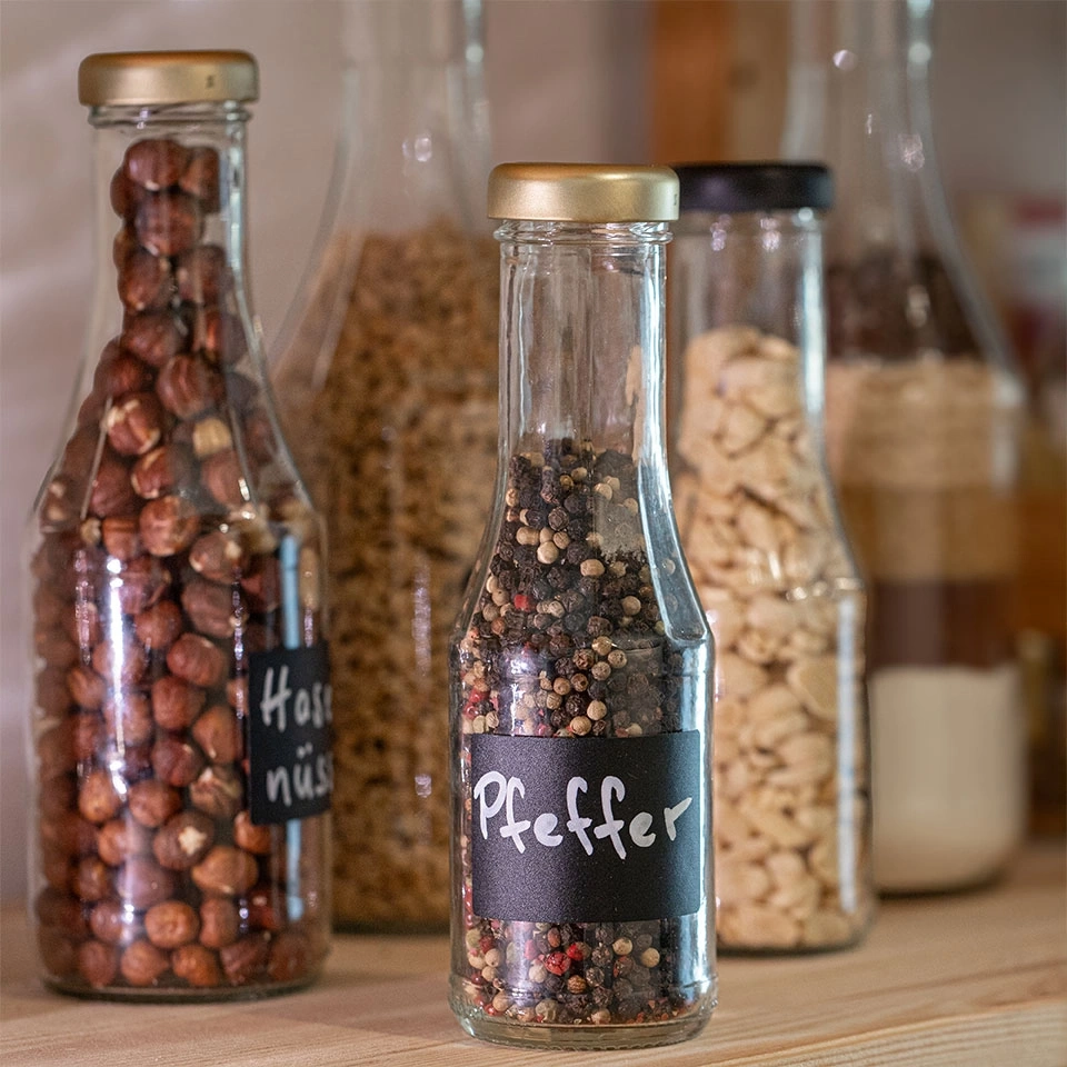 Glasflaschen mit Gewürzen und Nüssen, beschriftet mit 'Pfeffer' und 'Haselnüsse'.