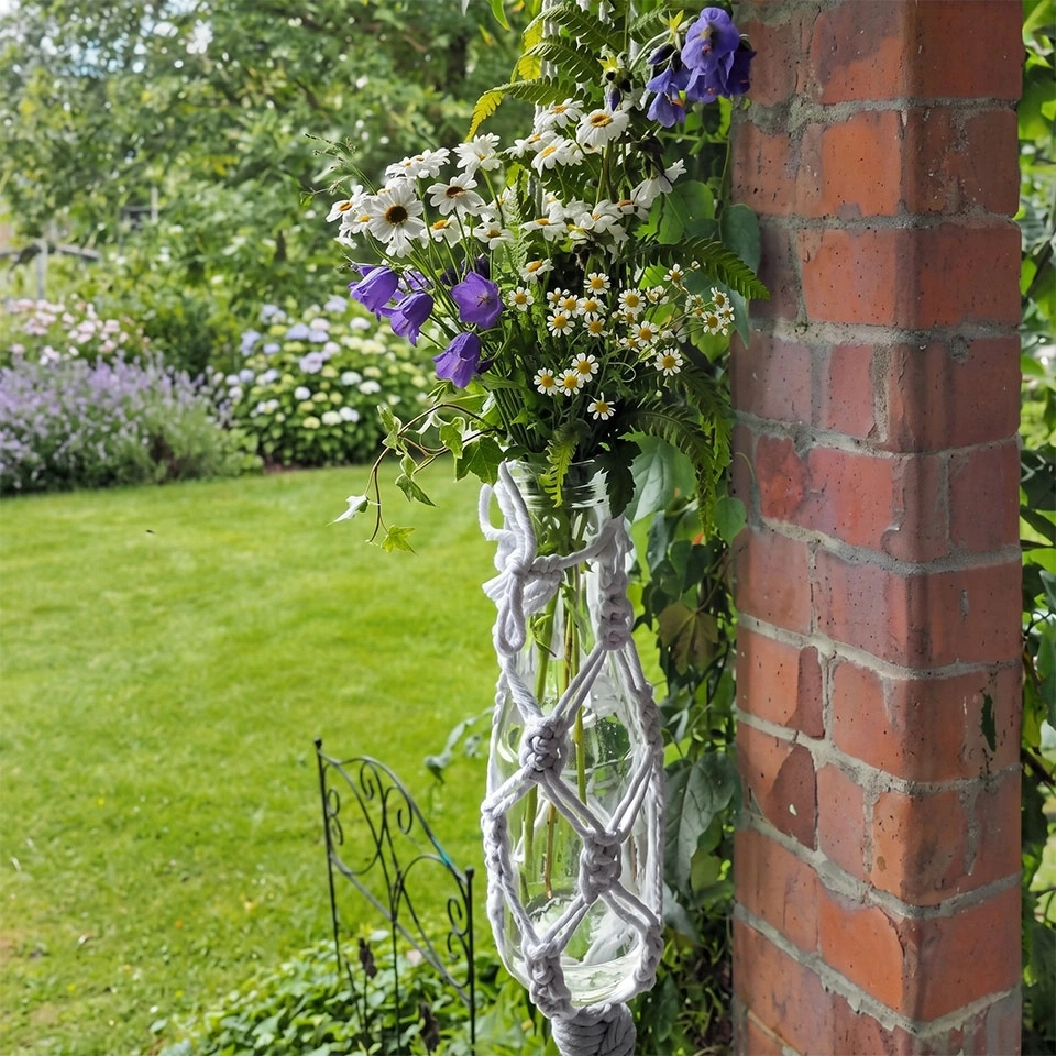 Ein Strauß Wildblumen und Glockenblumen hängt in einem Glasgefäß, das von einer Makramee-Halterung gehalten wird, die an einer Ziegelwand befestigt ist.