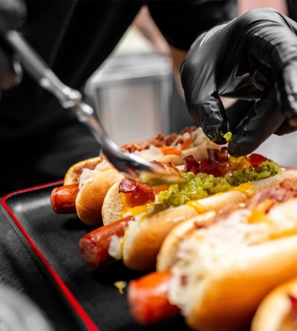 Ein Koch garniert Hot Dogs mit Gurken und Speck.