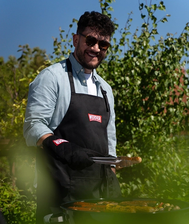 Ein Mann grillt mit einer Schürze und Handschuh im Freien.