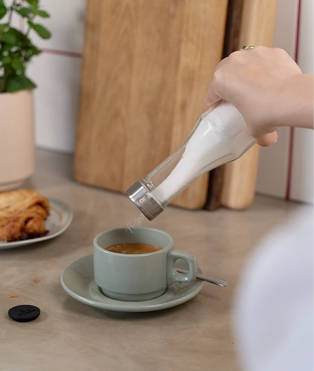 Eine Hand streut Zucker in eine Kaffeetasse über einem Gebäck.